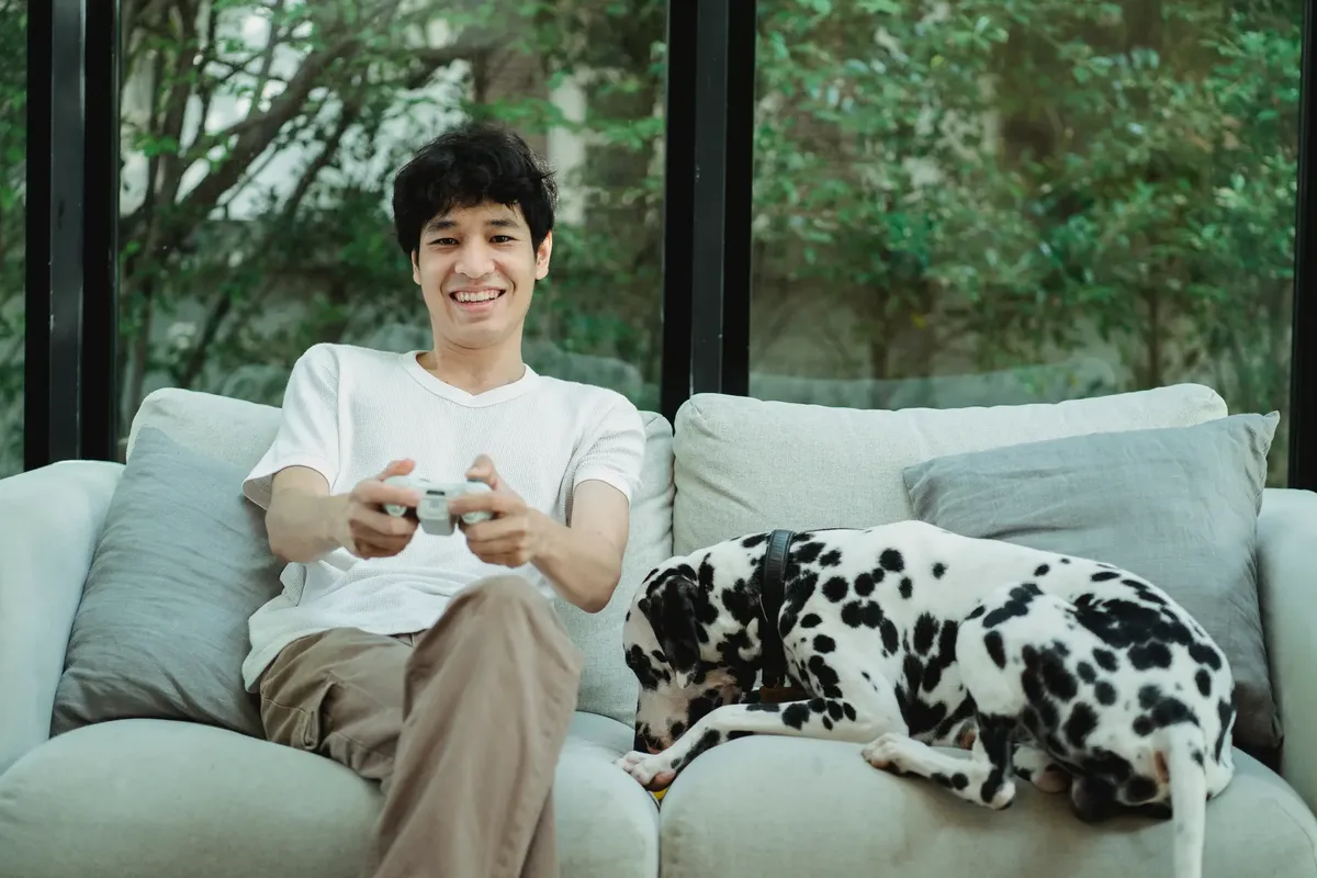 A young man enjoys video gaming on a sofa with his Dalmatian dog by his side.