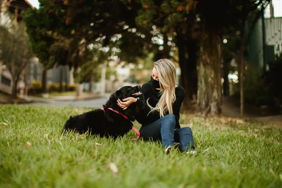 Serene female owner in protective mask stroking charming fluffy dog while sitting on lawn during COVID 19